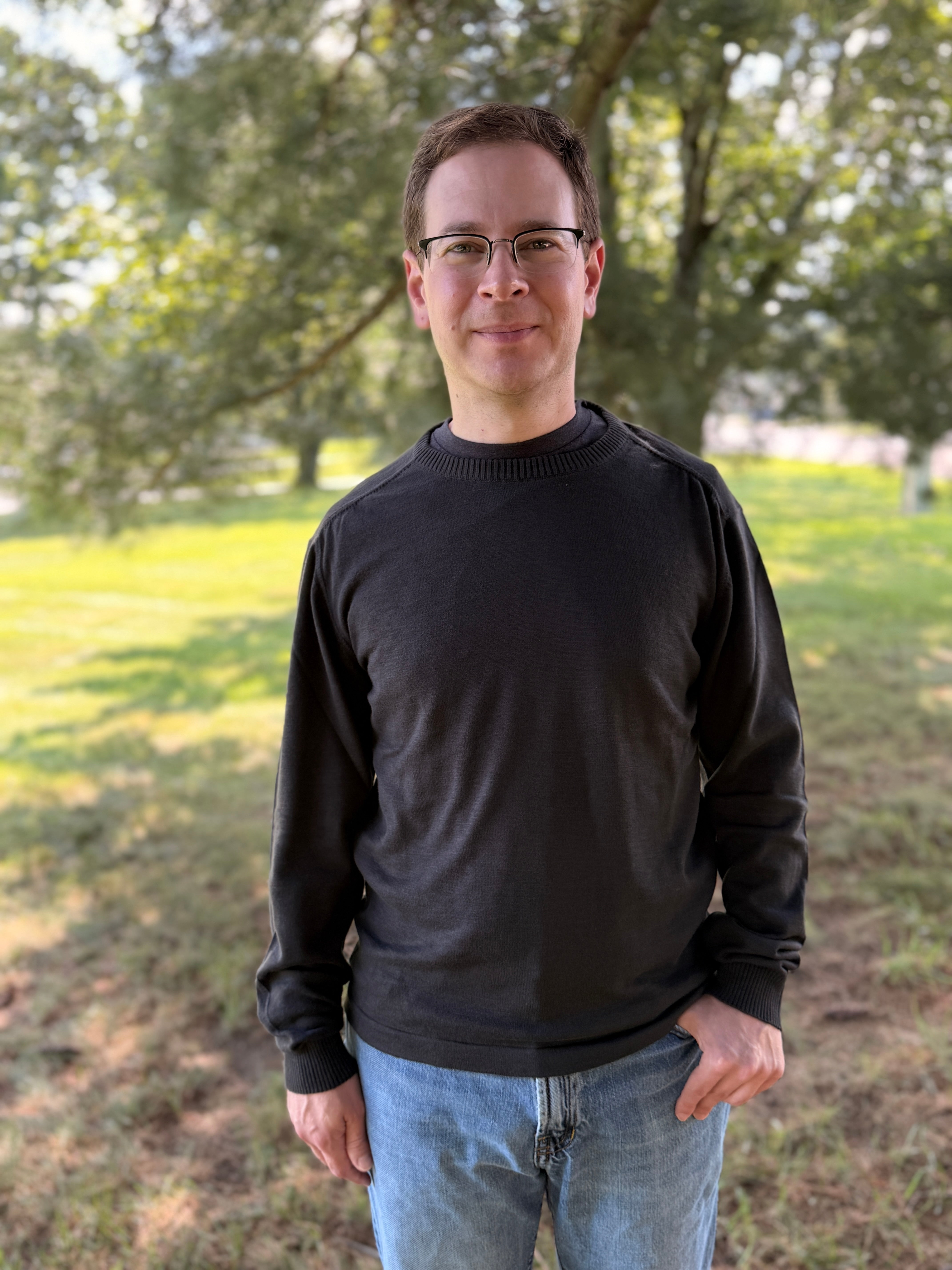 A man wearing a black long-sleeve crewneck sweater and blue jeans, standing outdoors in a grassy park area with trees in the background.