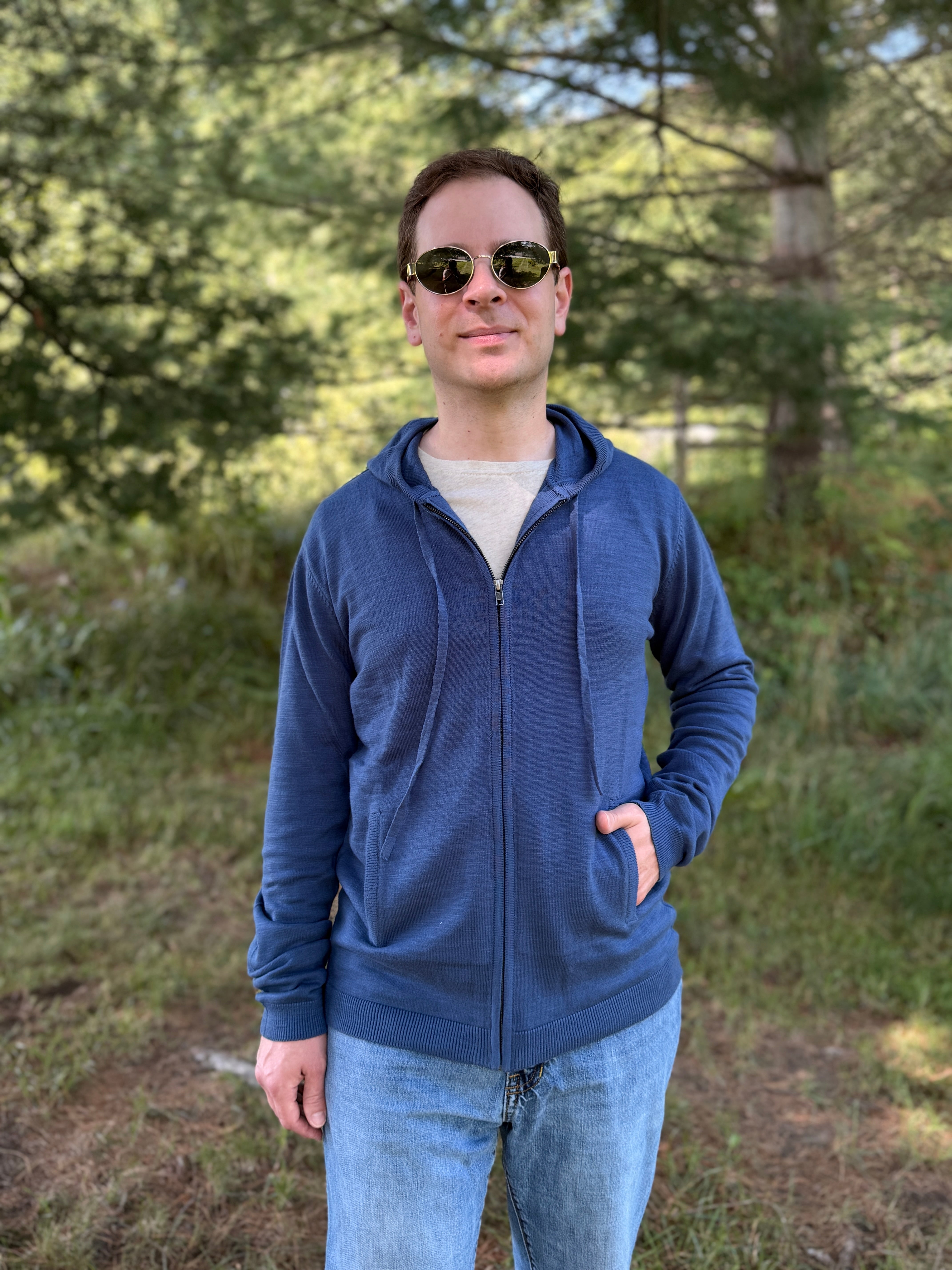 A man wearing a blue zip-up hoodie, light blue jeans, and sunglasses stands outdoors in a grassy, wooded area.