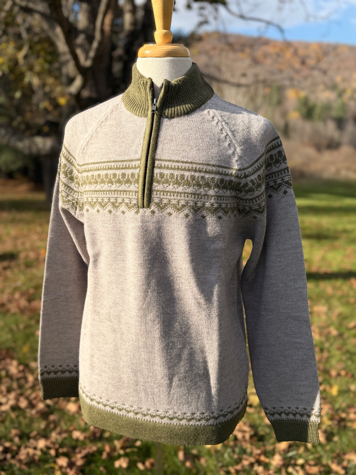 Grey quarter zip sweater with olive mock neck, sleeve and bottom rib trims, and pattern across chest and biceps. On mannequin against tree background.