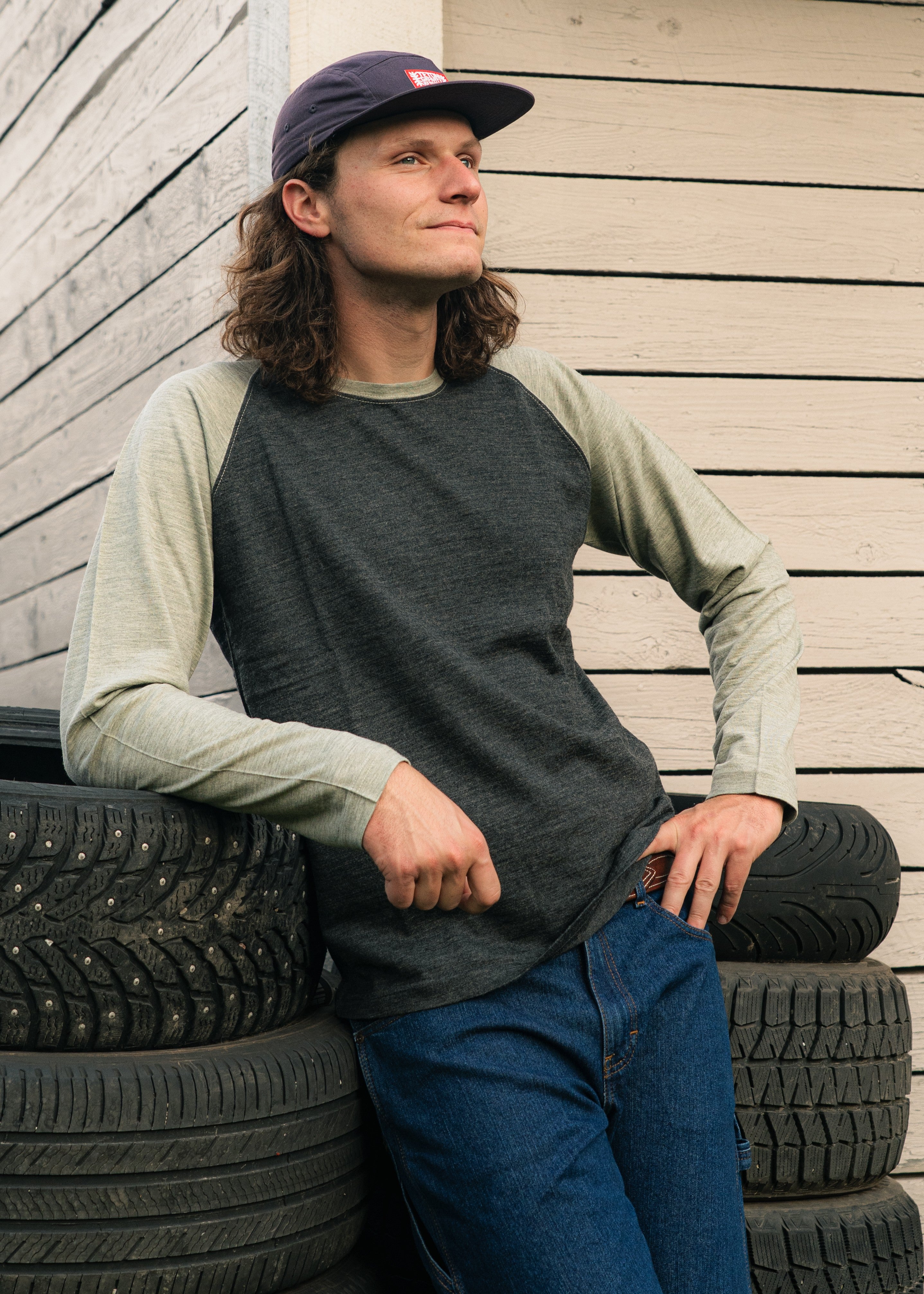 Man wearing a long-sleeve Home Team shirt and cap leaning against stacked tires