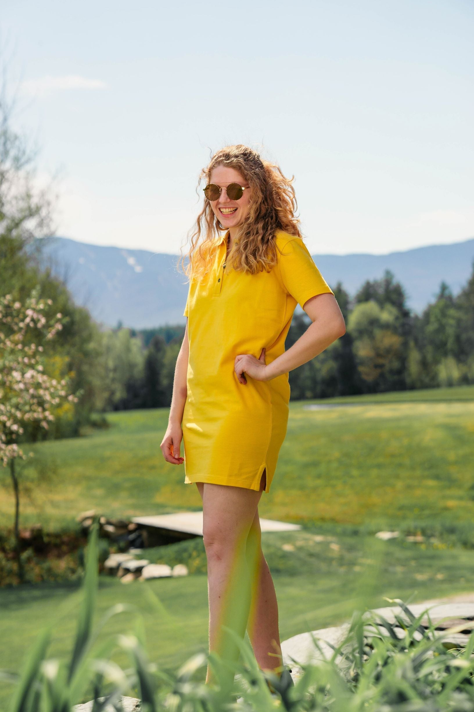 woman with sunglasses in yellow polo dress with short sleeves standing in front of grass