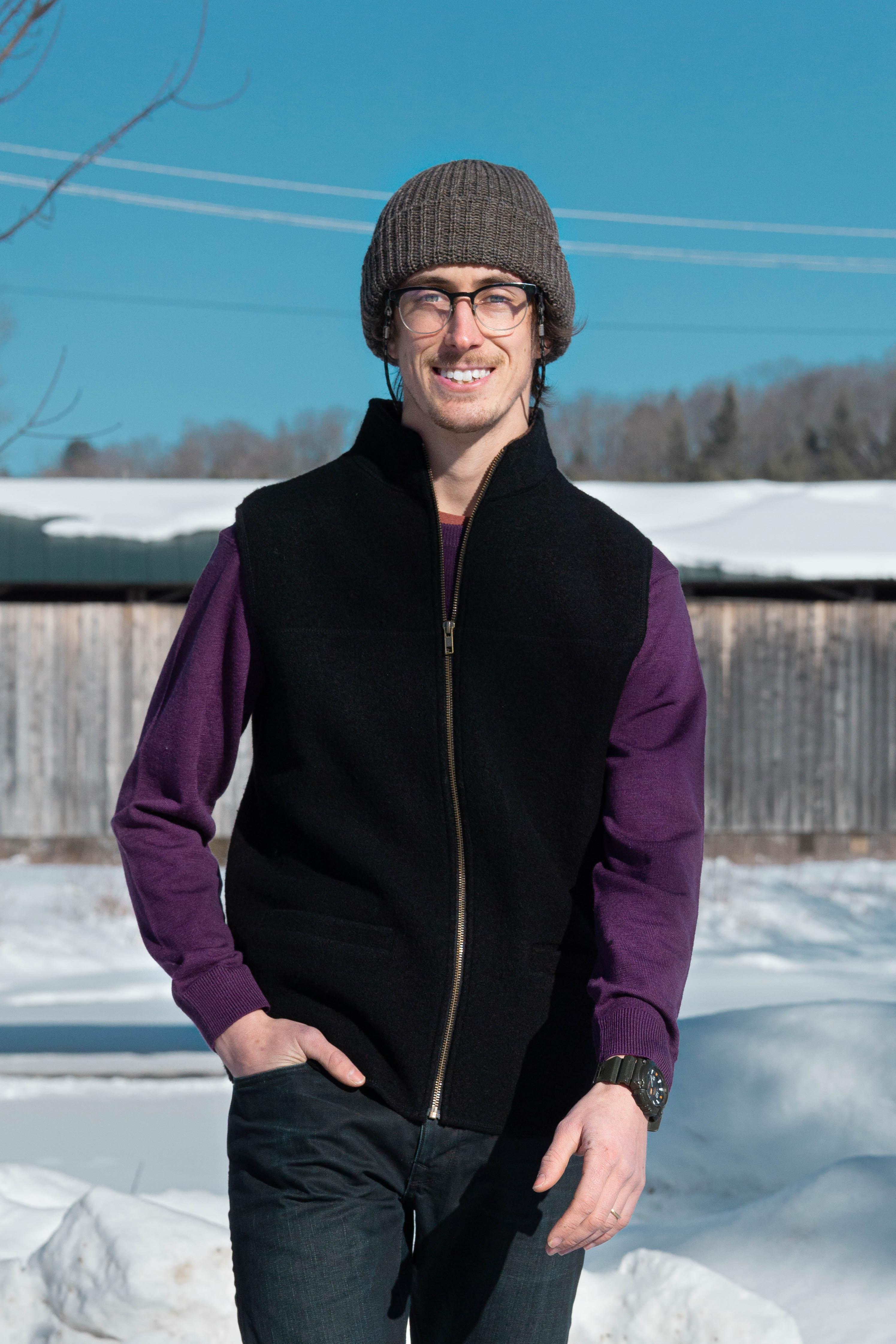 Man in boiled wool black vest over purple wool crewneck sweater with heathered grey wool knit hat in front of covered bridge