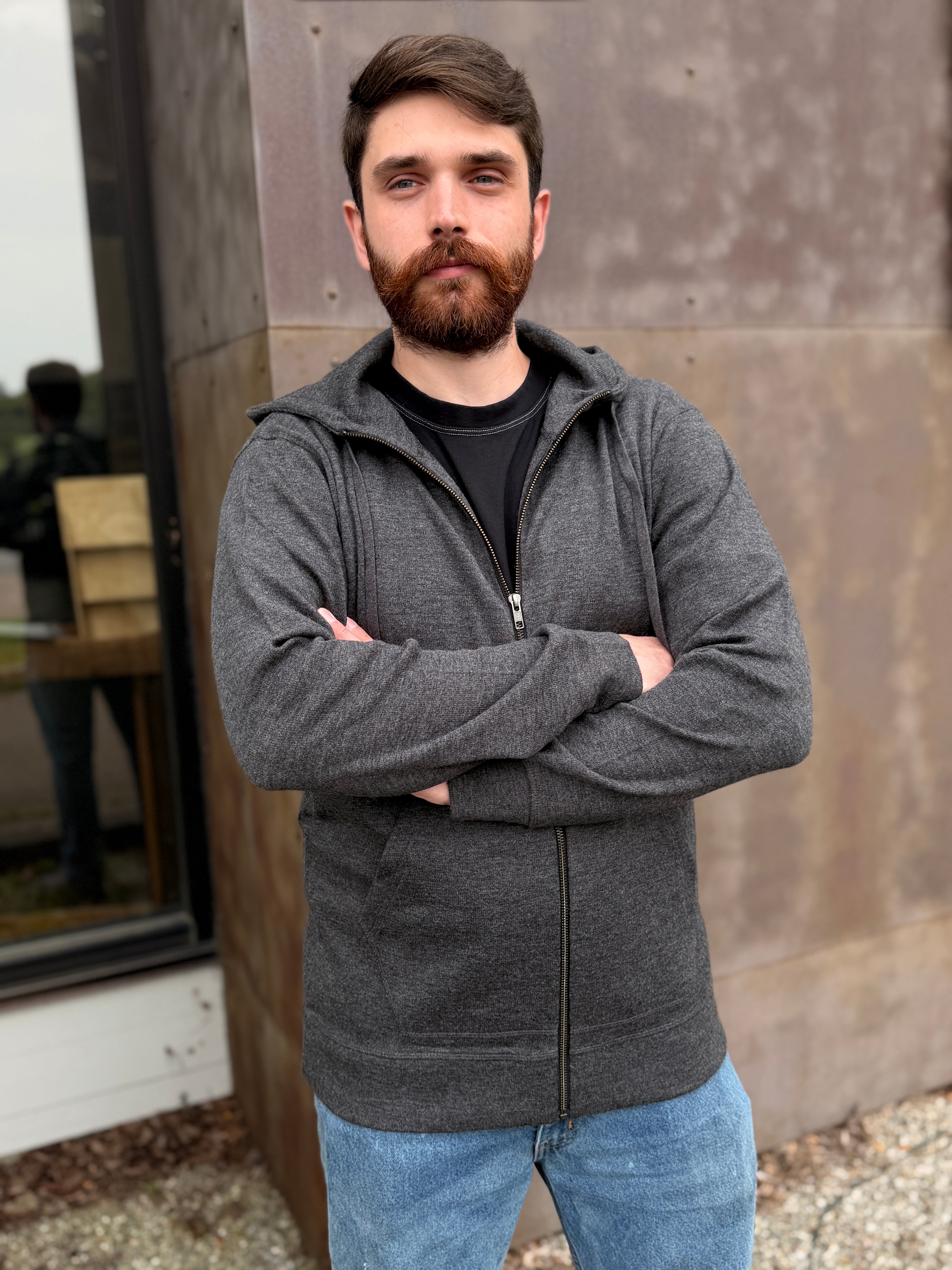 A man with a beard wearing a dark gray zip-up hoodie over a black t-shirt and blue jeans, standing with arms crossed in front of a rustic metal wall.