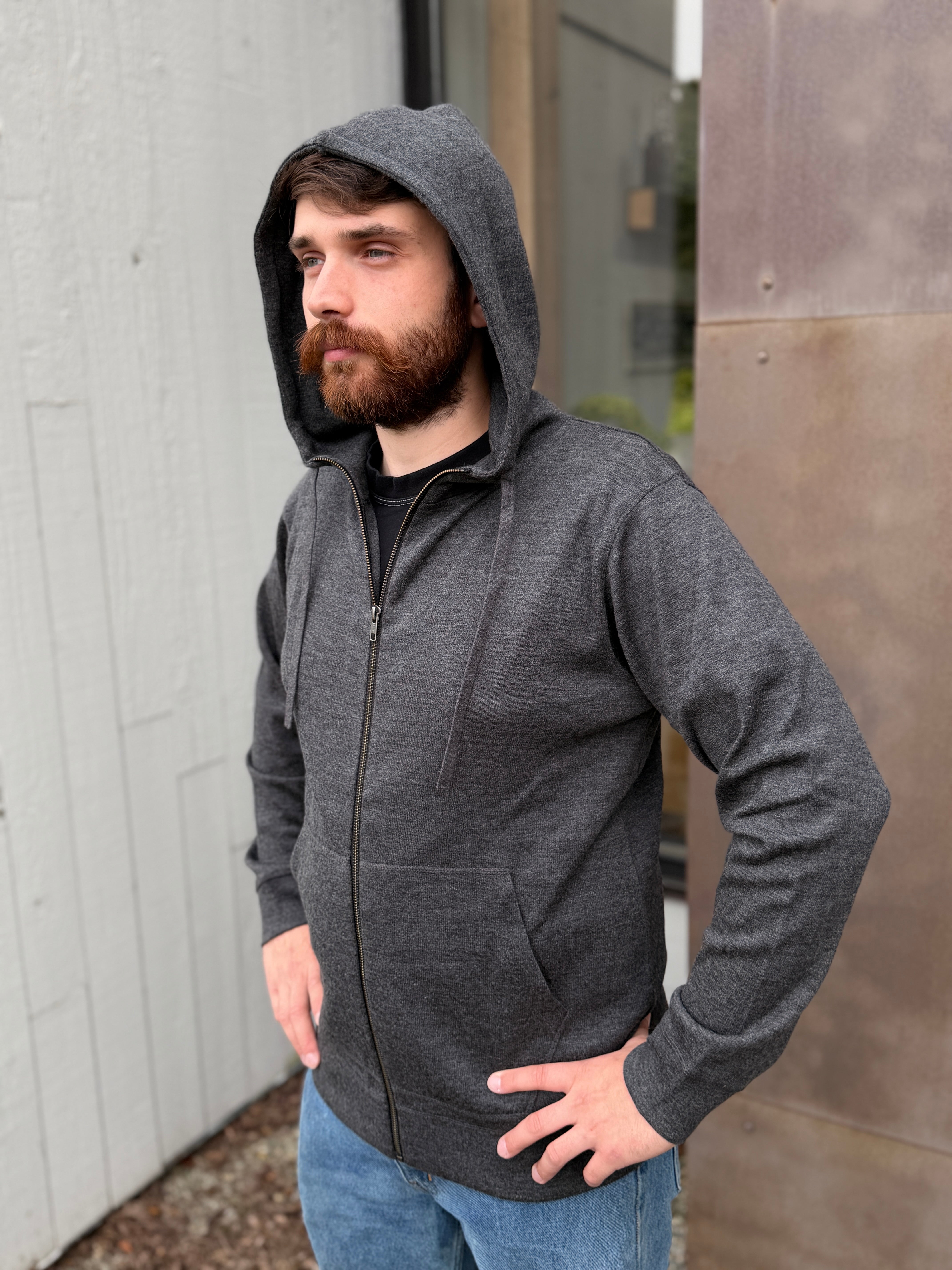 A man wearing a dark gray heather zip-up hoodie with a front pocket, standing outdoors against a modern building wall.