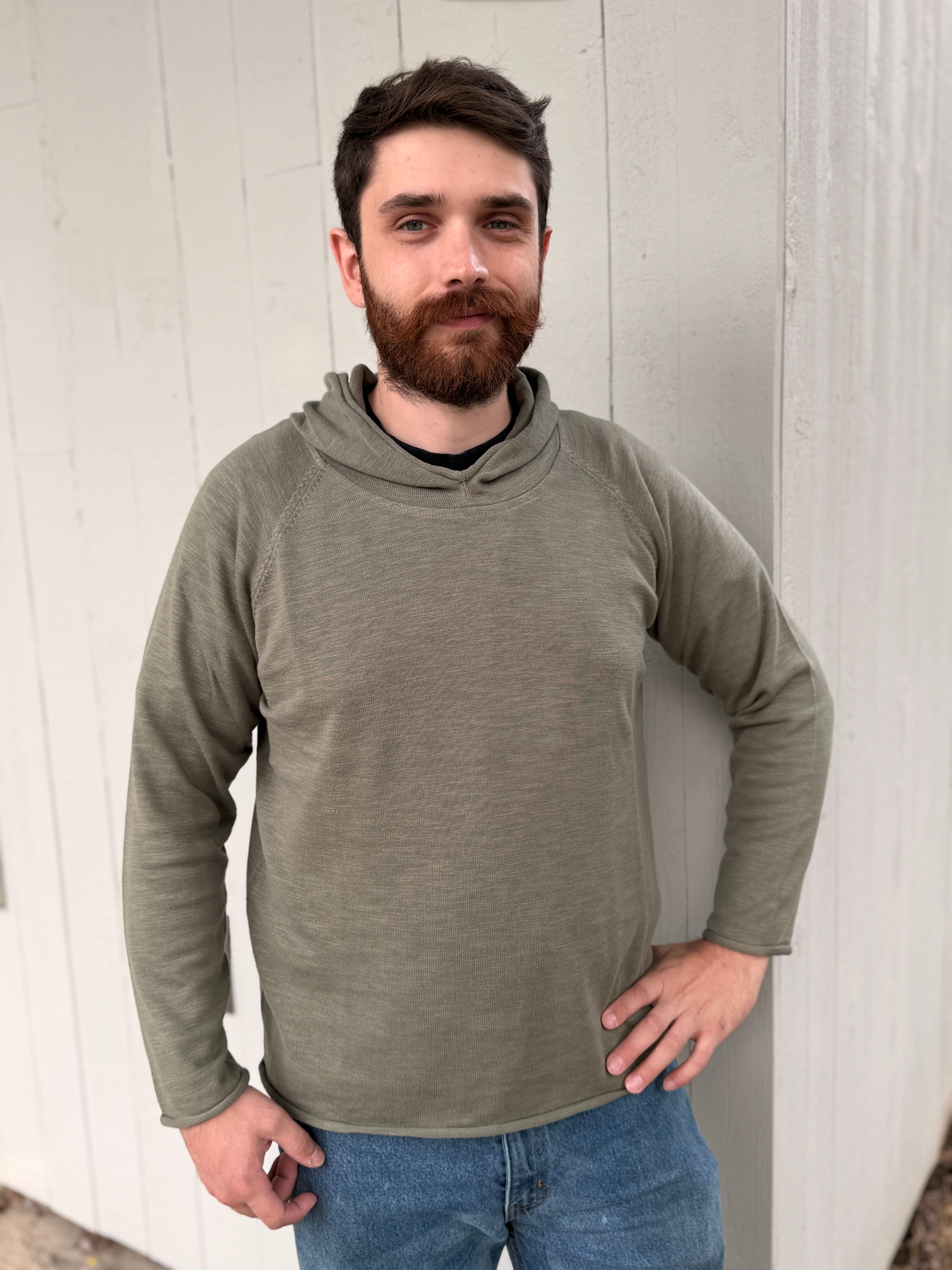 A man with a beard wearing an olive green long-sleeve hooded shirt and blue jeans, standing against a white wooden wall.