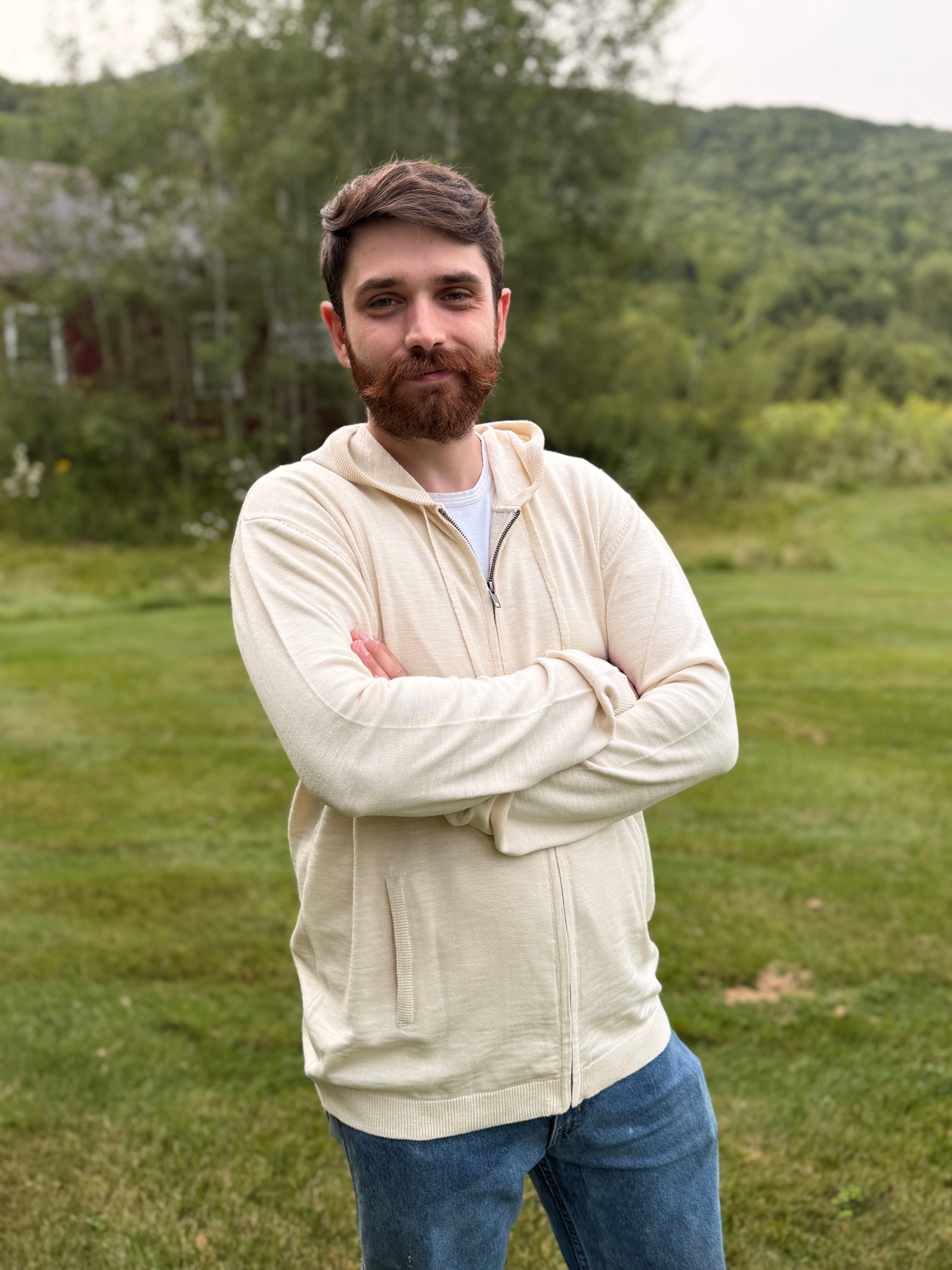 A man with a reddish beard stands outdoors on a grassy lawn with trees and hills in the background, wearing a cream-colored zip-up hoodie over a white t-shirt and blue jeans.