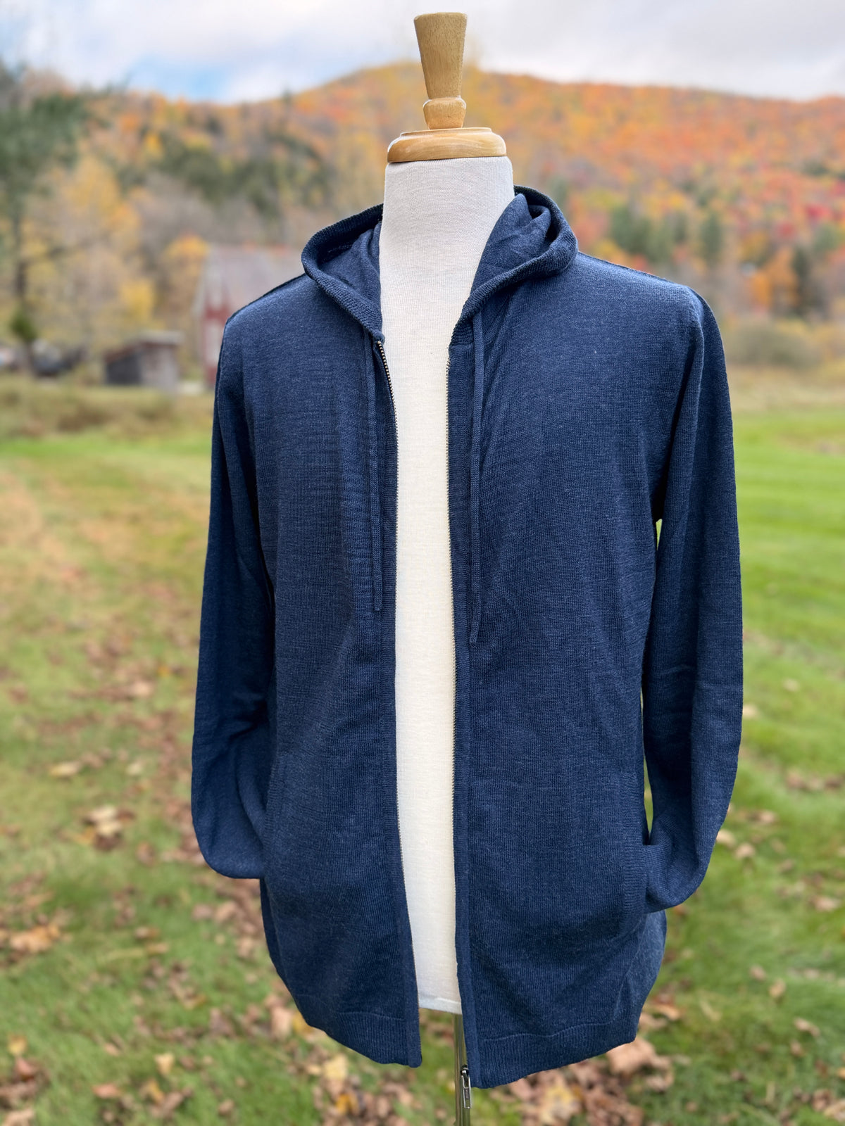 Unzipped blue hoodie on mannequin in grassy field with fall leaves behind it . Sleeves tucked in pockets as if a person has their hand in their pockets. 