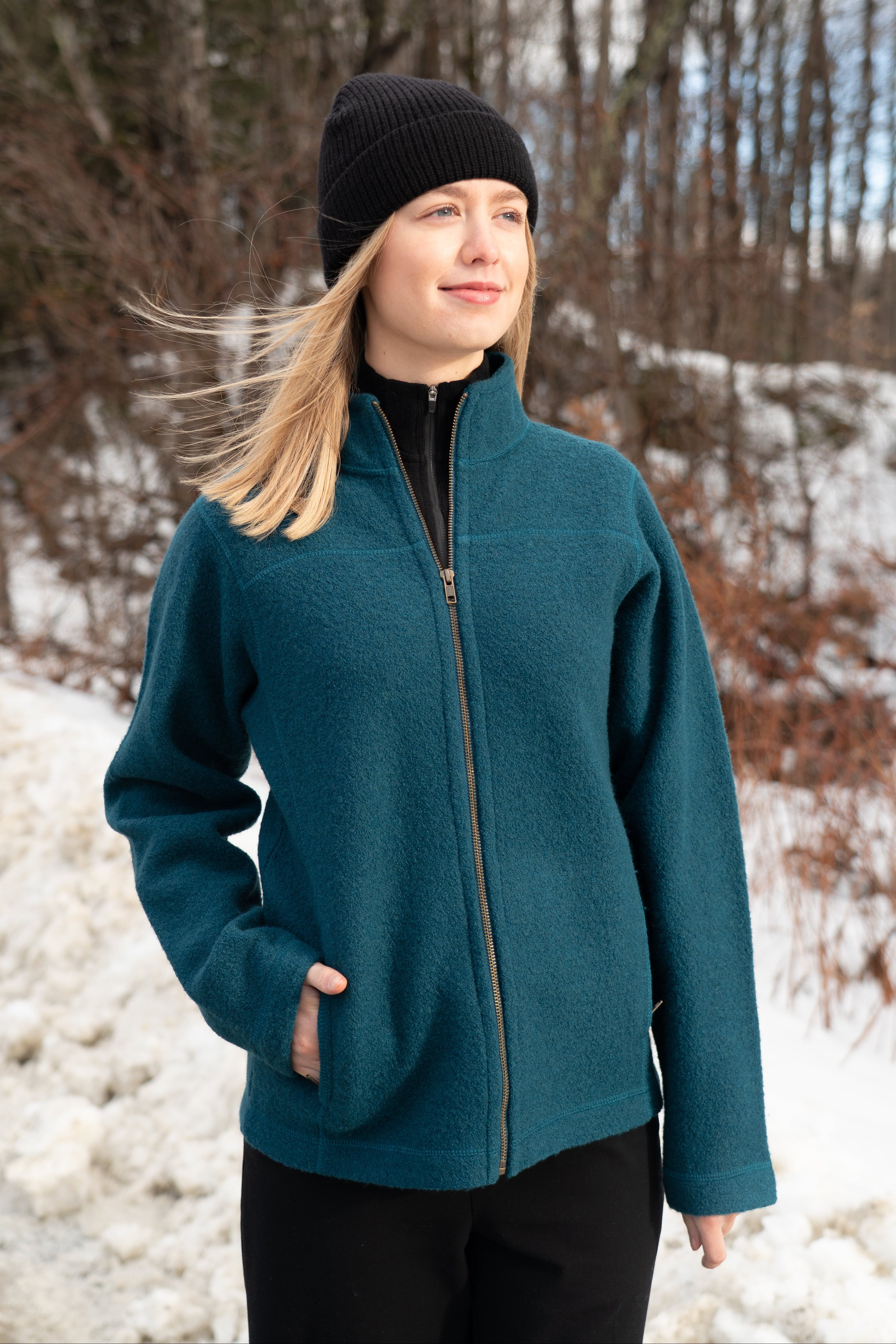 Person wearing a teal jacket and black beanie standing in a snowy landscape with bare trees.