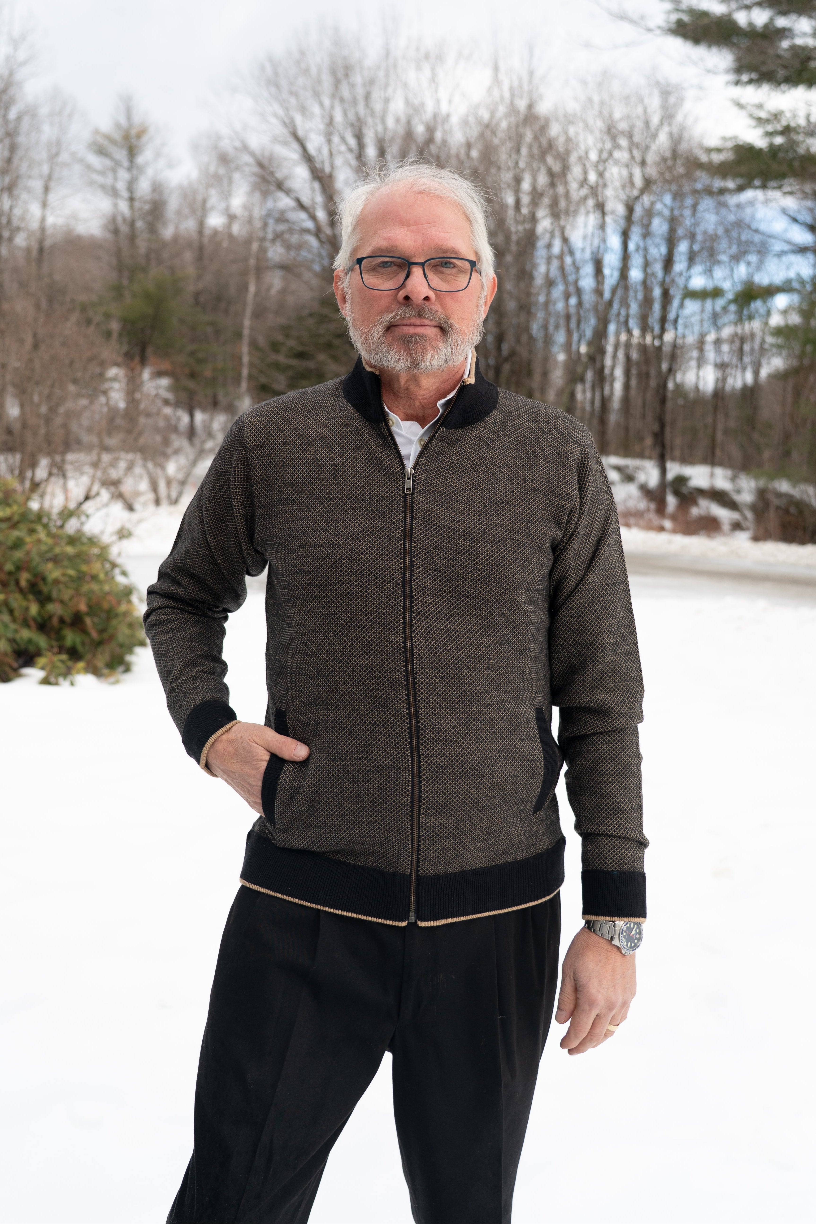 Man wearing a gray zip-up jacket and black pants standing in a snowy landscape with trees in the background.
